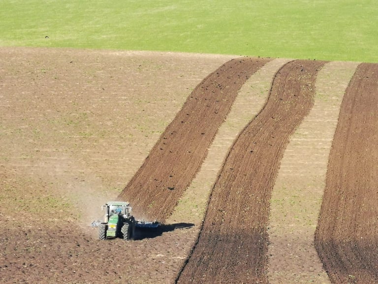 Tractor plowing a large field with alternating rows of brown and light-colored soil