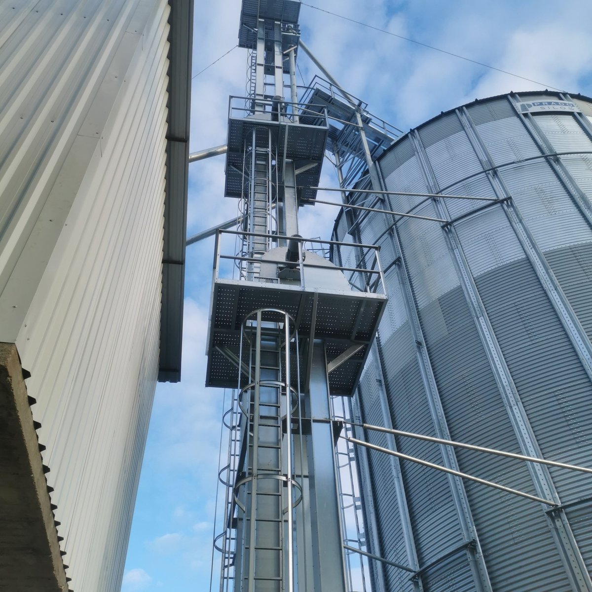 Industrial grain silo with metal staircase tower and corrugated metal buildings against blue sky