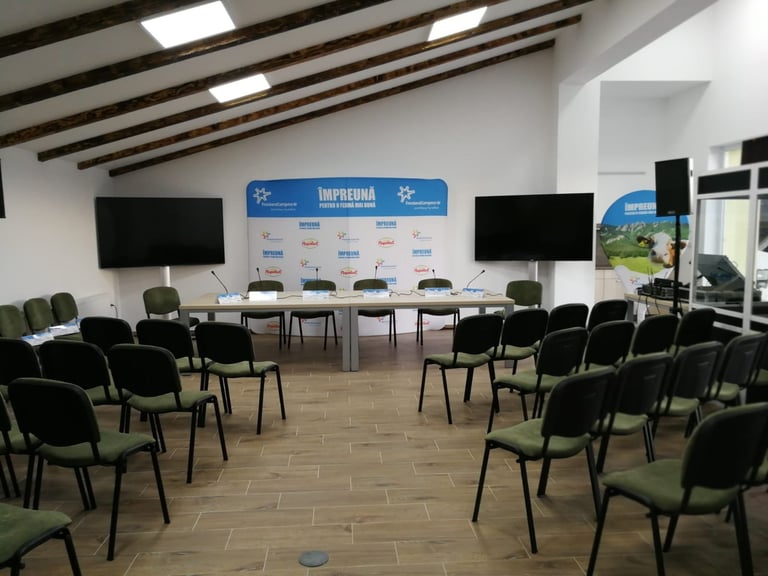 Empty conference room with black chairs arranged in rows facing a blue branded stage backdrop and two large screens
