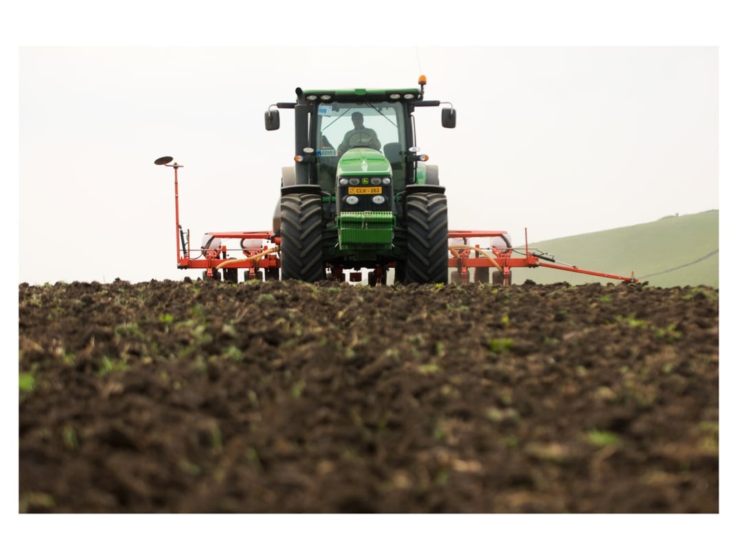 Green tractor with red attachment plowing brown soil field with hills in background